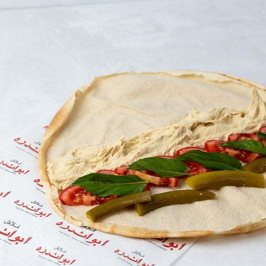 A large Hummus Sandwich on a flatbread with visible layers of hummus, tomato, cucumber, and mint leaves, served for you at Falafel Abou Andre