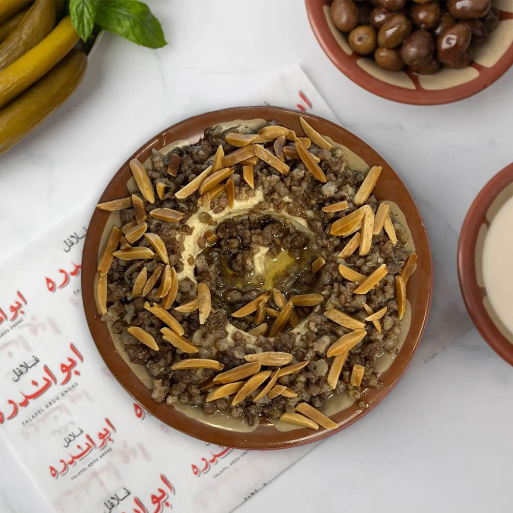 A plate of Lebanese Qawarma hummus with nuts, accompanied by pickled vegetables and olives, served with Lebanese bread.
